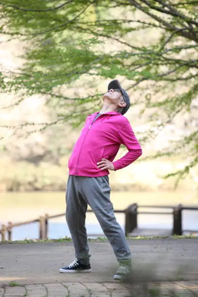 senior Japanese man in pink wear doing physical exercise standing back bend at the park