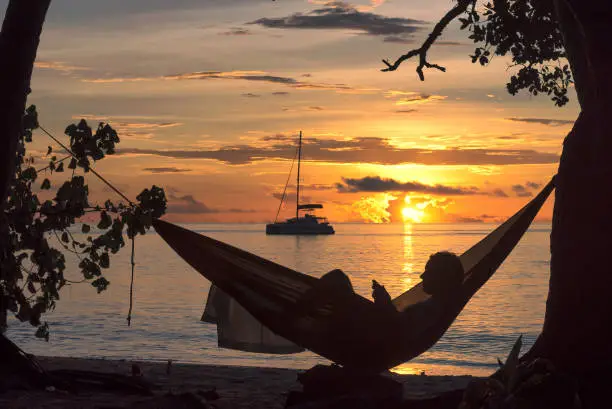 Photo of Beach vacations, silhouette of a man reading in hammock at sunset on tropical island.