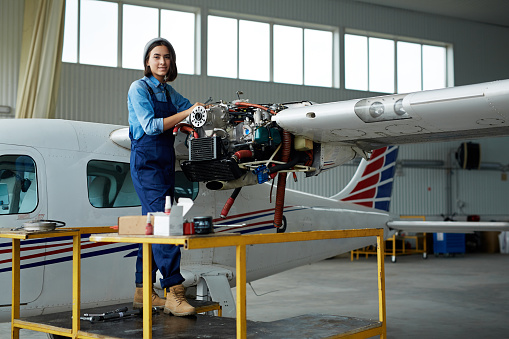 Portrait of modern young woman wearing overalls repairing jet plane turbine in hangar and smiling