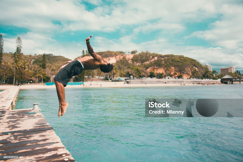 Backflip Pacific Islander jumping into the Water Noumea Beach New Caledonia Young pacific islander from the local kanak people tribe, the original inhabitants of New Caledonia jumping from wooden jetty with a backflip into the turquoise lagoon at the Beach of Noumea, New Caledonia. Real Native People Portrait. Noumea, New Caledonia, South West Pacific Ocean New Caledonia Stock Photo