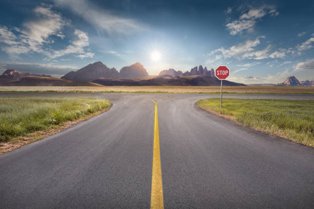 Decision for the right and a wrong way at intersection Crossing with stop sign towards the sun and mountain peaks with yellow line on asphalt. Success and vision concept. crossroad stock pictures, royalty-free photos & images
