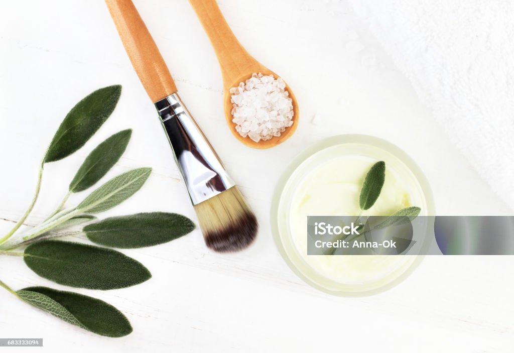 Herbal botanical facial mask with salvia. Ingredients for home spa top view Jar of cream, green plant leaves, sea salt, white wooden table background. Skin Care Stock Photo