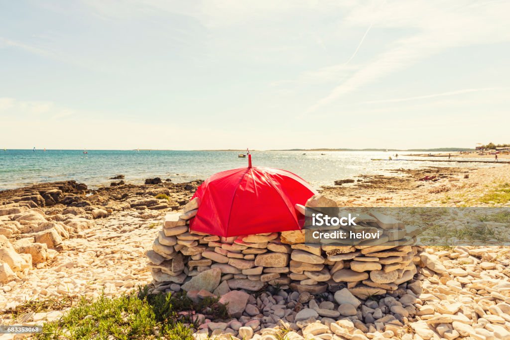 Red umbrella on the beach Stony beach and red umbrella covering stack of stones for a shadow. Bay of Water Stock Photo