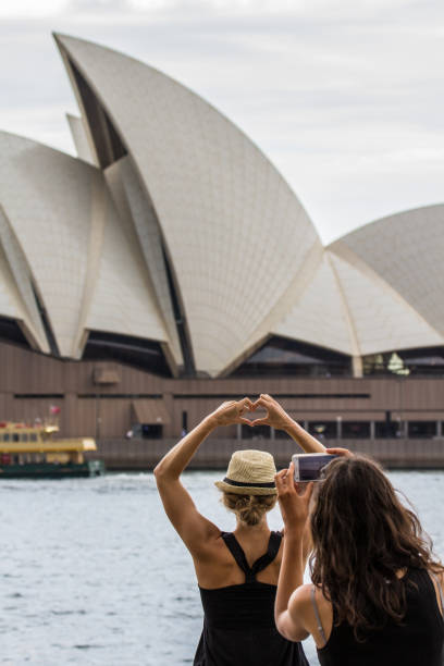 Sydney, NSW Australia – November 30th 2016 Sydney Opera House typical roof shape with cloudy sky and two persons taking a picture with hands forming a heard