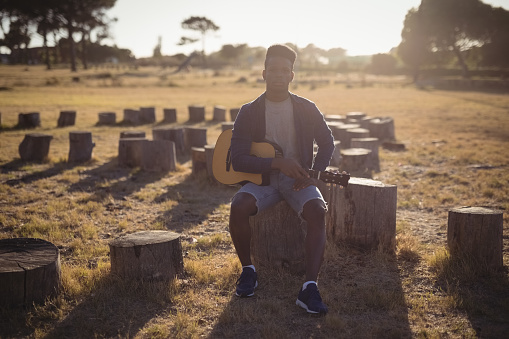 Full length of young man playing guitar while sitting on tree stump in forest