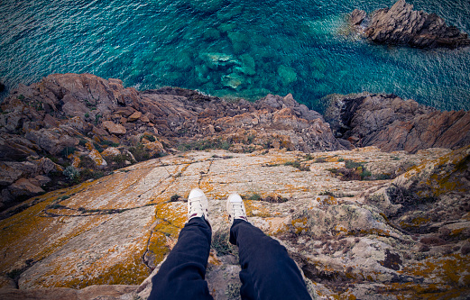 A man standing on a high cliff of an Italian coast with the Mediterranean sea Below him. Porto Cervo - Emerald Coast, Sardinia - Italy