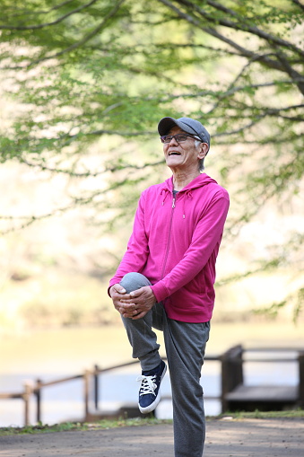 portrait of senior Japanese man in pink wear doing standing knee to chest stretch in the park