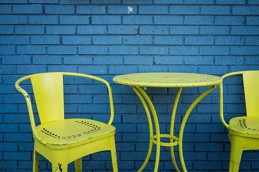 Brightly painted neon yellow metal patio chairs and table against a blue painted brick wall.