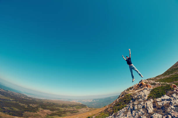 hombre joven en jeans y camisa negra sobre una roca de la montaña saltando en el cielo. - on top of business cliff businessman fotografías e imágenes de stock