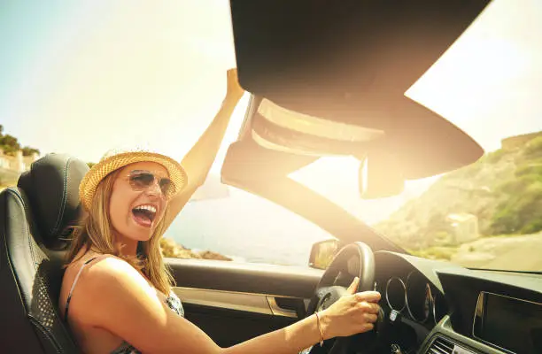 Single laughing or celebrating young woman with hat and sunglasses driving her convertible top automobile on bright sunny day near ocean