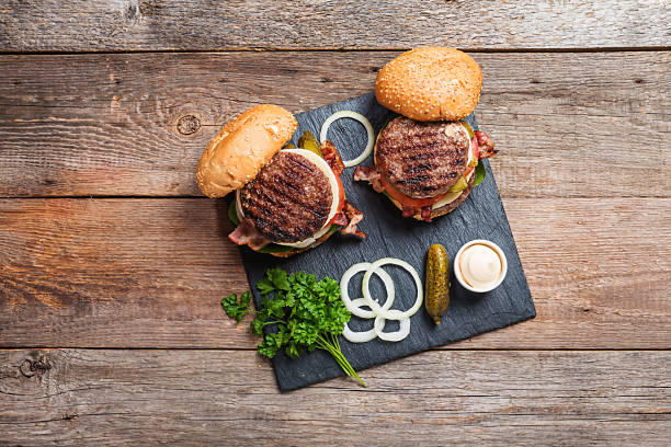 Fresh homemade burgers Two fresh homemade burgers on dark slate stone board, pickles, sauce and sliced onion over wooden background. Top view, copy space burger close up stock pictures, royalty-free photos & images