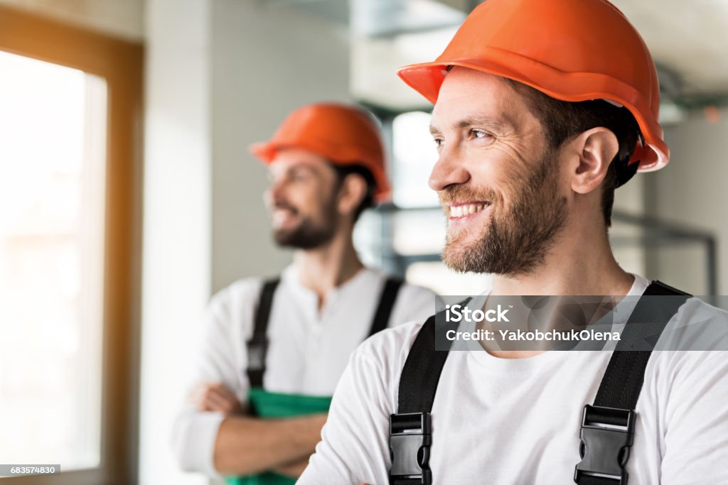 Hilarious smiling erectors in room Happy laughing builders are standing at quarters and looking aside with smile. They wearing orange helmet. Portrait Repairperson - Occupation Stock Photo