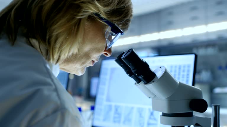 Senior Female Biologist Works on Her Computer and Looks at Materials under Microscope. She's in a Modern Laboratory.