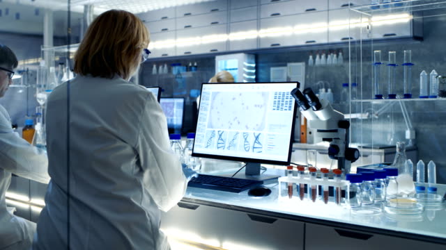 Senior Female Scientist Works with High Tech Equipment in a Modern Laboratory. Her Colleagues are Working Beside Her.