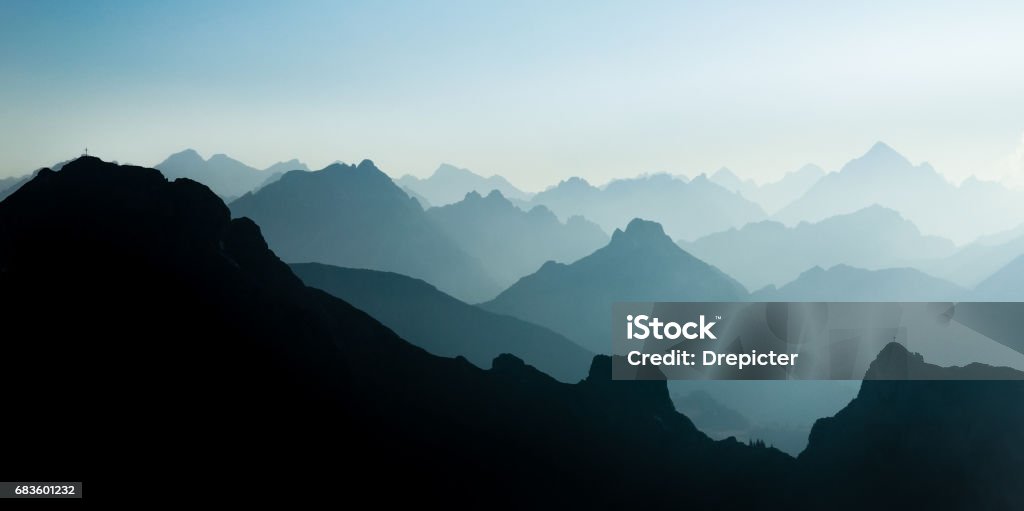 Spectacular blue and cyan mountain ranges silhouettes. Summit crosses visible. Blue and cyan mountain ranges silhouette with bright back light. Tirol, Austria. Summit crosses are visible from afar on some mountain peaks. Austria Stock Photo