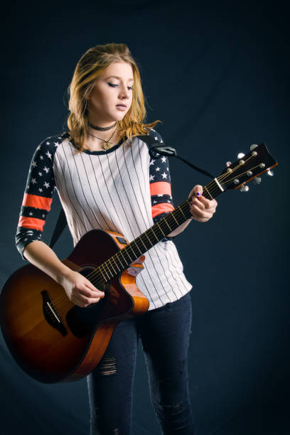 Confidence woman guitarist playing six string acoustic guitar and looking at camera, standing full length. Young adult female musician with glasses is dressed in red leather jacket, jeans and sneakers