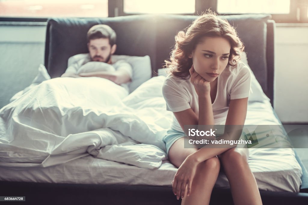 Couple in bedroom We have a problem. Young upset girl sitting on the edge of the bed, against her boyfriend, lying in bed. Couple - Relationship Stock Photo