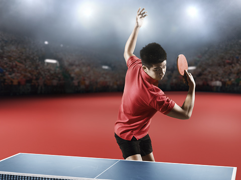 Young asian ping pong player playing table tennis game. He is holding a red racket. He is very concentrated. Behind him, the stadium with the fans in the defocus.