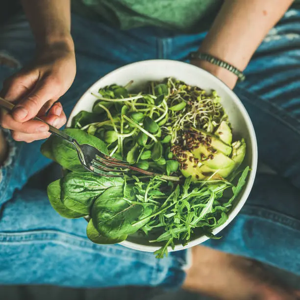 Green vegan breakfast meal in bowl with spinach, arugula, avocado, seeds and sprouts. Girl in jeans holding fork with knees and hands visible, top view, square crop. Clean eating, dieting food concept