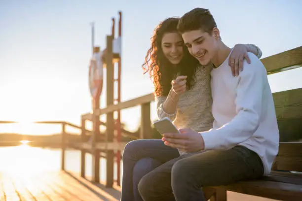 Photo of Happy teenage couple sitting outdoors in nature and using mobile phone with sunset and lens flare