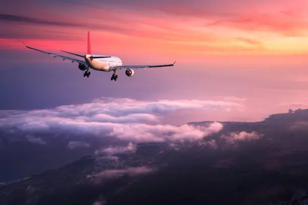 Photo of Airplane. Landscape with big white passenger airplane is flying in the red sky over the clouds at colorful sunset. Journey. Passenger airliner is landing at dusk. Business trip. Commercial plane
