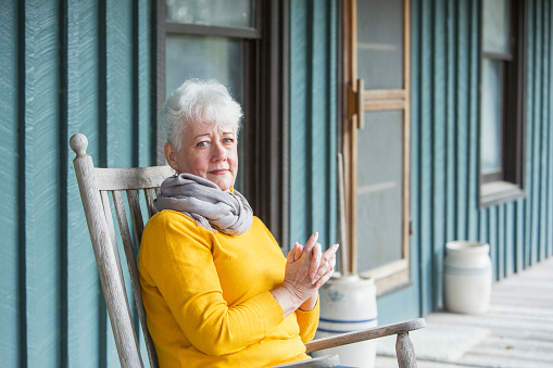 A senior senior woman in her 60s sitting in a rocking chair on a porch, looking at the camera, hands clasped.