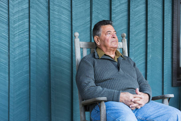 uomo anziano serio seduto su una sedia a dondolo sotto il portico - men senior adult serious depression foto e immagini stock
