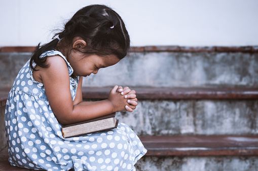 Cute asian little girl closed her eyes and  folded her hand in prayer on a Holy Bible for faith concept in vintage color tone