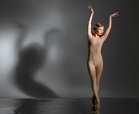 Beautiful young woman modern ballet dancer dressed in skin colored ballet leotard. She performs the ballet pose. The ballerina is standing tiptoe. Her hands are raised over head. Her eyes are closed. Shooting in studio on foil covered floor on gray background, the shadow of the dancer on wall