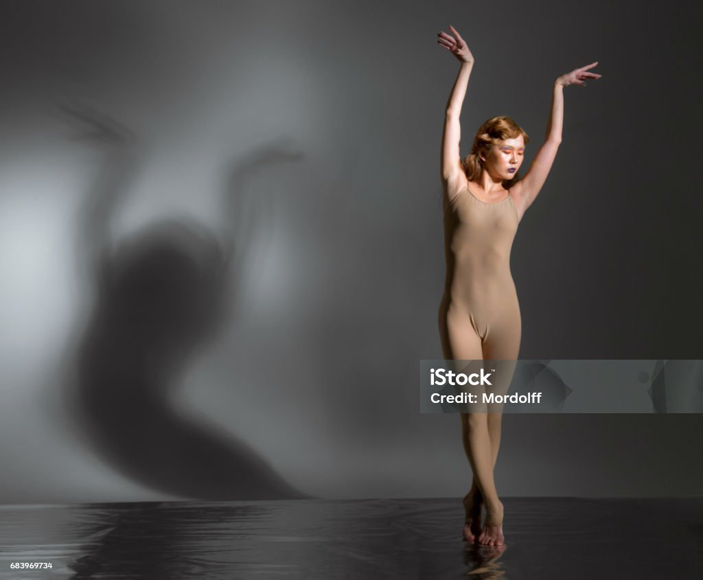 Young Japanese Ballerina Beautiful young woman modern ballet dancer dressed in skin colored ballet leotard. She performs the ballet pose. The ballerina is standing tiptoe. Her hands are raised over head. Her eyes are closed. Shooting in studio on foil covered floor on gray background, the shadow of the dancer on wall Activity Stock Photo