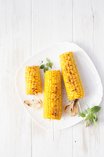 The cob fried corn on a white plate with sliced grilled onions and herbs on white wooden table.