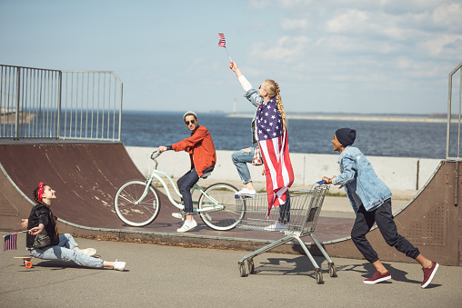 Teenagers group with skateboard, bicycle and shopping cart having fun together and waving american flags at skateboard park