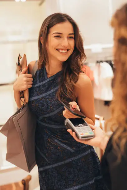 Beautiful elegant woman paying via smart phone at a clothing store.