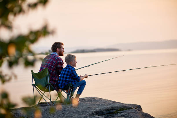 Father and Son Enjoying Fishing Together Side view portrait of father and son sitting together on rocks fishing with rods in calm lake waters with landscape of setting sun, both wearing checkered shirts, shot from behind tree son stock pictures, royalty-free photos & images