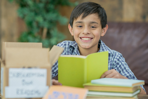 A monthly subscription box of books and bookmarks sits on a table in front of a hispanic boy who is reading and smiling in a living room.