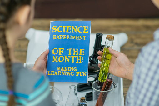 A boy and girl open a monthly science experiment subscription box together. They pull out educational materials like test tubes, beakers, and a microscope.