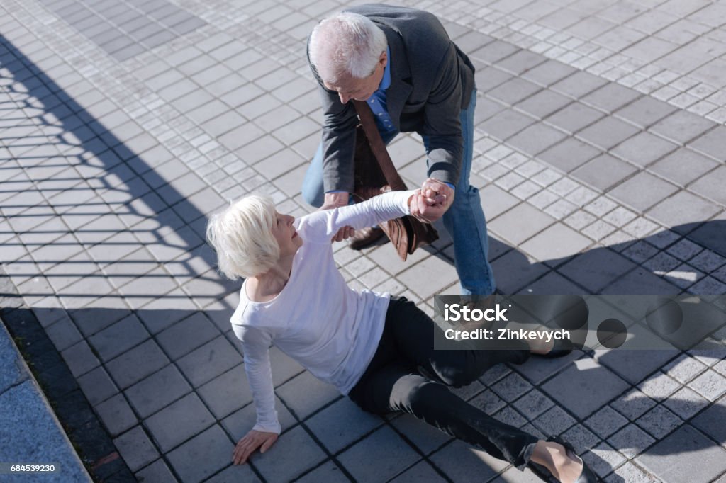 Pleasant woman tumbling over outdoors Do not afraid of falling down. Funny white-haired slim lady catching foot and receipting of relief from aged man who smiling Falling Stock Photo