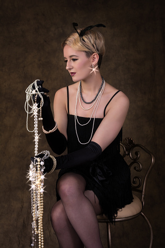 Vertical studio shot on dark backdrop of 17 year old blonde model in 1920s flapper dress, black with fringe. Seated holding handfulls of sparkly jewelry with 6 pointed stars on highlights.