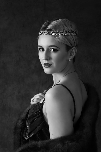Vertical B&W studio shot of 17 year old blonde model in vintage style dress with interesting neck line, and head band. Head and shoulders looking back over shoulder at camera. Slight smile.