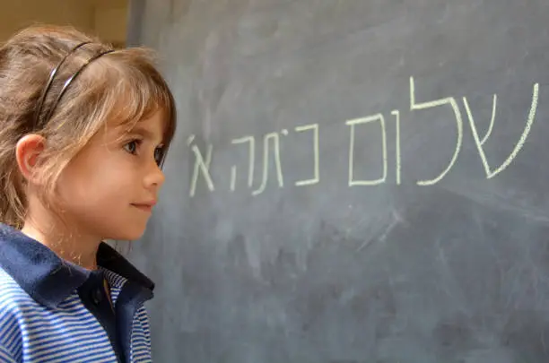 Little girl (Age 5-6) reads Hello First Grade greetings in Hebrew (Shalom Kita Alef) on a chalkboard in Israeli primary school at the beginning of the school year. Education concept photo