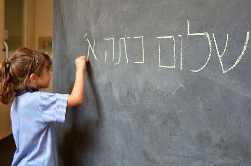 Little girl (Age 5-6) writes Hello First Grade greetings in Hebrew (Shalom Kita Alef) on a chalkboard in Israeli primary school at the beginning of the school year. Education concept photo