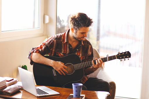 Young man playing an acoustic guitar in a brightly lit room. On a desk in front of him is a laptop, coffe mug, and some books. His gaze is directed to his hands, while he tries to play a song.