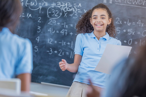 Confident African American female STEM student gives an oral report in front of her classmates. She is standing in front of a chalkboard. Math problems are written on the chalk board.