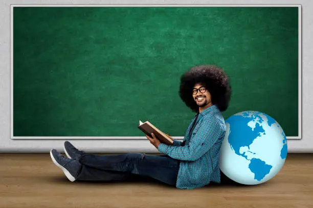 Afro college student holding a book while sitting with a globe and smiling at the camera