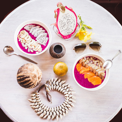 Healthy and tasty fresh breakfast on vintage round table served with sliced mango and dragon fruit, fresh papaya, coconut, smoothie. Lay flat in tropical colors with sunglasses, flowers, bijou and food, view from above