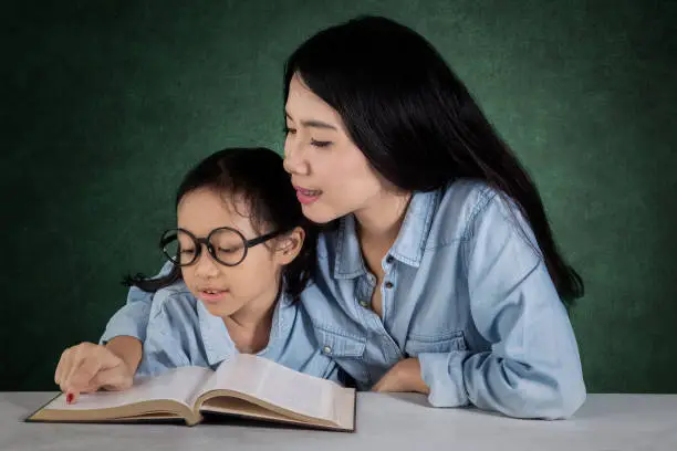 Portrait of a cute student reading a book with a female teacher in the classroom