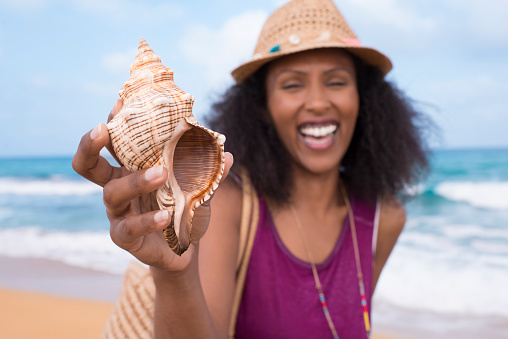 Happy tourist woman showing shell of conch. Selective focus on seashell. Relaxed Afro-American woman on sea beach. Summer holiday concept image.