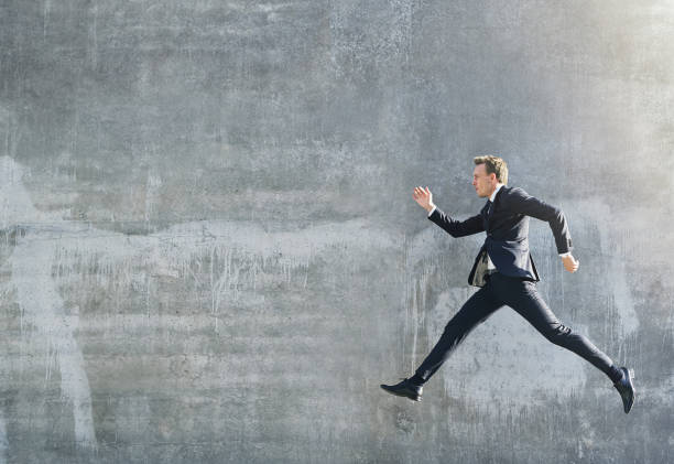 Side view of man in suit caught in jump making pose of walking on grey wall