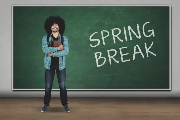 Young man holding a book while standing with spring break word on chalkboard