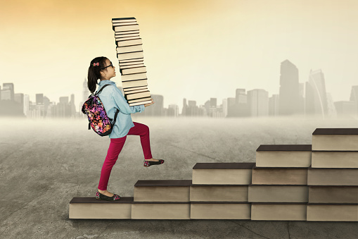 Female elementary school student carrying a pile of books and walking upward on books shaped a stair